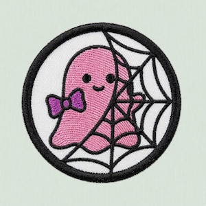 以下が含まれることがあります： 紫色の蝶ネクタイをしたピンク色の幽霊が描かれた円形の刺繍ワッペン。背景は白で、黒いクモの巣のデザインが施されています。円形のワッペンの縁は、太くしっかりとした黒いステッチで囲まれています。