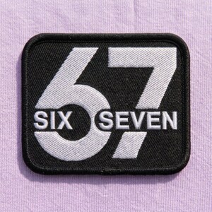 Puede incluir: Un parche rectangular negro bordado con esquinas redondeadas que presenta el número 67 en blanco grande. La palabra SIX está bordada en blanco dentro del número 6, y la palabra SEVEN está bordada en blanco dentro del número 7.
