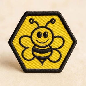 May include: A hexagonal embroidered patch featuring a yellow background with a black border. The design depicts a cartoon bee with black and yellow stripes, large eyes, and antennae, centered within the hexagon shape to resemble a honeycomb cell.