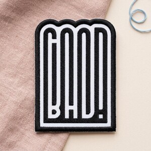 May include: A rectangular embroidered patch with a black border and white background. The patch features the word GOOD stacked vertically above the word BAD, both in bold black sans-serif lettering. The text includes an exclamation point at the end.