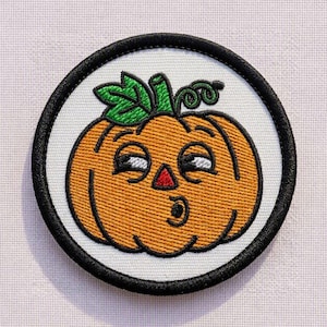 May include: A circular embroidered patch featuring an orange pumpkin with a green stem and curly vine. The pumpkin has a white face with black embroidered eyes, a small red triangular nose, and a black circular mouth on a white background.
