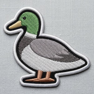 Op de afbeelding: Een geborduurde opstrijkbare patch in de vorm van een wilde eend. De eend heeft een groene kop, een beige snavel, een grijs lichaam en een witte borst met bruine poten. Het ontwerp is omlijnd met een dikke witte rand met zwarte afwerking.