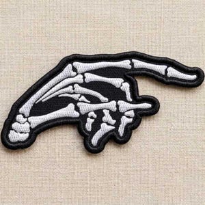 May include: An embroidered patch featuring a white skeleton hand on a black background. The hand is positioned with the index finger extended to point horizontally to the right, while the remaining fingers are curled inward toward the palm.