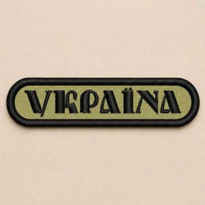 May include: A rectangular embroidered patch with rounded corners featuring a black border and a muted olive green background. The text "УКРАЇНА" is stitched in bold black capital letters across the center of the patch in a stylized font.