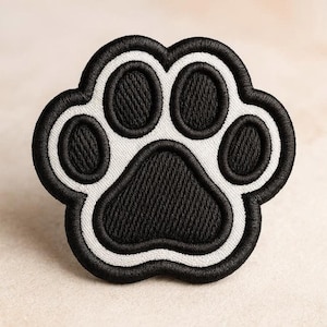 Pode incluir: Um emblema bordado em forma de pegada de animal. O design apresenta uma almofada central grande e quatro almofadas menores em fio preto, delineadas por uma borda branca espessa e um acabamento final costurado em preto.