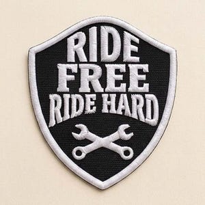 May include: A shield-shaped embroidered patch with a black background and white border. The text RIDE FREE RIDE HARD is stitched in white capital letters. Below the text, two white wrenches are crossed in an X shape, also embroidered in white.
