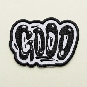 May include: An embroidered patch featuring the word GOOD in a bold, black, bubble-style graffiti font. The letters are outlined in black against a white background, with small white highlights inside the black lettering to create a glossy effect.