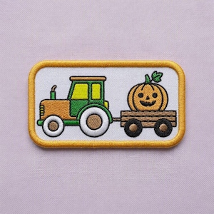 May include: A rectangular embroidered patch featuring a yellow and green tractor pulling a wooden wagon with a smiling orange jack-o'-lantern pumpkin. The design is set against a white background and framed by a thick, golden-yellow border.