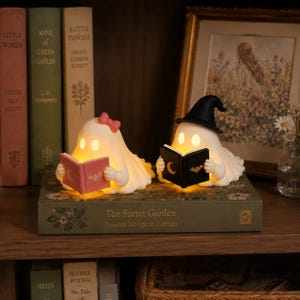 May include: Two white ghost figurines illuminated from within, positioned on a book titled The Secret Garden. One ghost wears a pink bow and reads a pink book with a moon and bat icon, while the other wears a black witch hat and reads a black book.