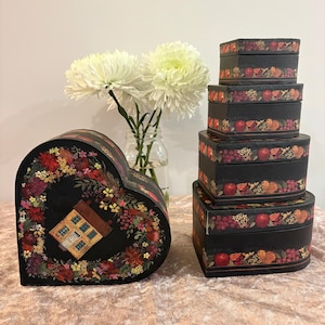 Könnte beinhalten: Ein Set aus schwarzen herzförmigen Schachteln mit einem bunten Blumen- und Fruchtmuster am Rand. Eine Schachtel zeigt in der Mitte die Abbildung eines kleinen Hauses. Die Schachteln sind in verschiedenen Größen gestapelt, daneben steht eine Glasvase.