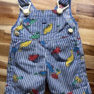 May include: Blue and white striped short overalls featuring a colorful dinosaur and palm tree print. The garment includes white plastic buttons on the sides and shoulder straps, with a small square logo patch labeled CPK on the front chest pocket.