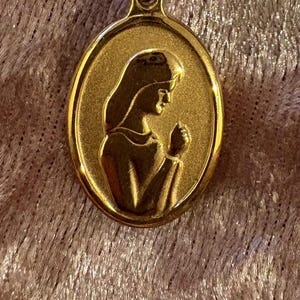 May include: A gold-toned oval pendant featuring a raised relief design of a person in profile with hands clasped together in a prayer-like gesture. The pendant has a smooth, polished border and a textured, matte-finish central background area.