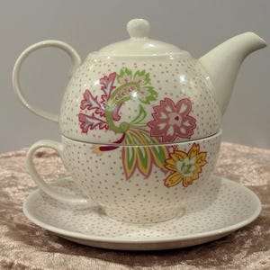 Könnte beinhalten: Ein weißes Keramik-Teeservice für eine Person, bestehend aus einer Teekanne, einer Teetasse und einer Untertasse. Das Set zeigt ein zartes Muster aus kleinen rosa Punkten und ein lebhaftes Blumenmuster in Rosa-, Gelb- und Grüntönen.