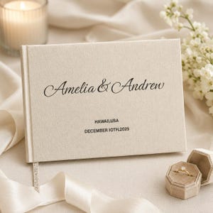 Puede incluir: Un libro de visitas rectangular cubierto de lino beige con el texto impreso en negro Amelia & Andrew, seguido de la ubicación Hawaii, USA y la fecha DECEMBER 10TH, 2025. Una pequeña cinta de tela marca páginas cuelga del borde inferior.