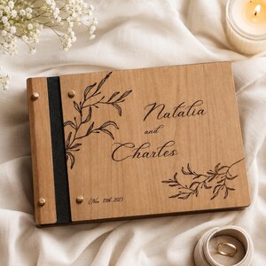 Puede incluir: Un álbum de fotos rectangular de madera con un lomo oscuro y tornillos de encuadernación de metal dorado. La cubierta presenta ilustraciones botánicas grabadas con láser, los nombres Natalia y Charles, y la fecha Nov 11th 2023 en cursiva.
