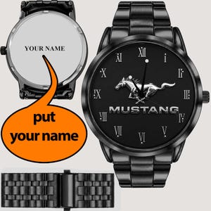 Op de afbeelding: Een zwart polshorloge met een metalen schakelband, voorzien van een zwarte wijzerplaat met een zilverkleurig Mustang-paardembleem, de tekst MUSTANG en Romeinse cijfers. De achterkant toont de tekst YOUR NAME, met een apart beeld van de sluiting.