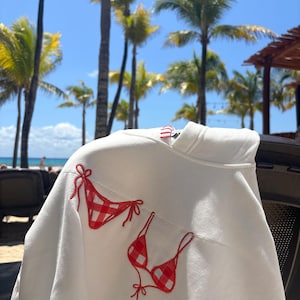 May include: A white hooded sweatshirt featuring an embroidered design of a red and white gingham bikini. The embroidery includes a bikini top and bottom with red thread detailing for the straps and ties, positioned on the front of the garment.