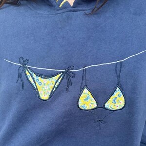 May include: A navy blue hooded sweatshirt featuring an embroidered design of a bikini set hanging on a white clothesline. The bikini pieces are yellow with a blue floral and butterfly pattern, outlined in dark blue thread with tie details.