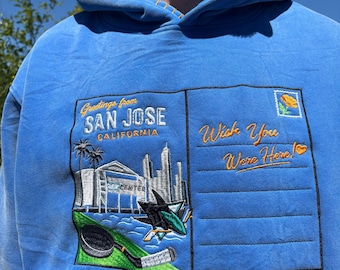 May include: A bright blue hooded sweatshirt featuring an embroidered postcard design on the front. The graphic includes the text "Greetings from SAN JOSE CALIFORNIA" and "Wish You Were Here!" with a city skyline, a hockey stick, and a puck.