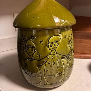 May include: A glossy olive green ceramic canister with a matching lid, featuring embossed illustrations of three figures in pointed hats holding hands, accented by small star shapes and geometric patterns around the cylindrical body.