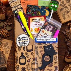 May include: A brown paper-wrapped book package featuring a botanical print, a bookmark labeled Crows, Corn & Moonlight, a reading list card, a Twinings tea bag, a neon yellow pen, a Frankenstein lollipop, and a black cat thumb page holder.