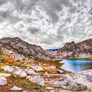 May include: A panoramic landscape featuring a deep blue alpine lake surrounded by rugged, light-colored rocky mountains. The foreground is covered in scattered boulders and patches of vibrant orange and yellow autumn vegetation under a cloudy sky.