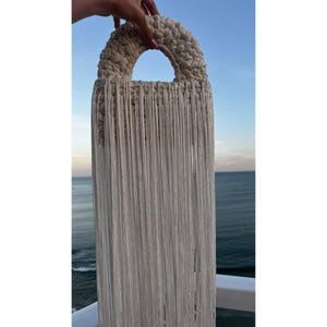Pode incluir: Uma bolsa de mão semicircular na cor creme com uma alça superior de crochê grossa e uma franja longa e densa feita de cordões brancos verticais finos que pendem da base da estrutura da alça em uma linha reta e uniforme.