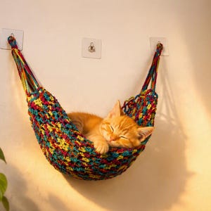 Pode incluir: Uma rede de gato em crochê multicolorida com uma mistura vibrante de fios vermelhos, amarelos, azuis e verdes. A rede está suspensa na parede por dois ganchos adesivos transparentes, criando um berço suspenso para um gatinho laranja a dormir.