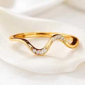 Puede incluir: Un anillo dorado con forma de onda que presenta una banda lisa y pulida con una sección central curva. La parte curva está decorada con una hilera de cinco pequeñas piedras claras de talla redonda engastadas en el metal, creando un diseño fino.