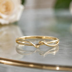 Può includere: Un anello in oro lucido caratterizzato da un delicato design intrecciato. La fascia è composta da due sottili fili d'oro lisci che si incrociano al centro, creando una silhouette continua, fluida e a forma di anello aperto.