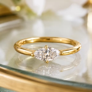 Puede incluir: Un anillo de oro con una sola piedra preciosa transparente de talla marquesa engastada horizontalmente en el centro. La banda es lisa y pulida, mostrando un diseño sencillo y limpio que resalta la forma alargada de la piedra central.