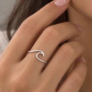 May include: A thin, polished silver-coloured ring featuring a single, continuous wire design shaped into a stylised ocean wave. The band is slender and minimalist, wrapping around the finger with a smooth, reflective metallic finish.