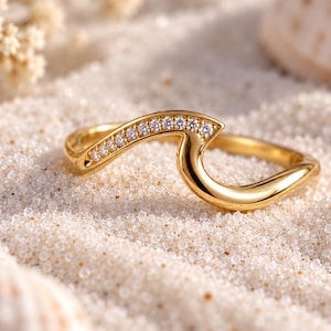 Puede incluir: Un anillo de oro con forma de ola oceánica estilizada, que presenta una banda pulida y lisa en un lado y una hilera de pequeñas piedras redondas y transparentes incrustadas a lo largo de la cresta de la ola, con un acabado metálico brillante.