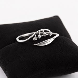 Op de afbeelding: Een zilverkleurige ring met een delicaat, open ontwerp. De band is gevormd als een slanke stengel die rondbuigt, versierd met drie kleine, bungelende klokvormige bloesems en een enkel langwerpig, glad blad aan het uiteinde.