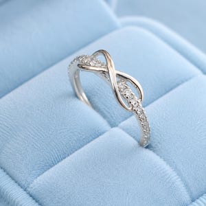 Puede incluir: Un anillo de tono plateado que presenta un diseño de símbolo de infinito en la parte superior. La banda y el bucle del infinito están incrustados con pequeñas piedras claras y brillantes. El anillo tiene un acabado metálico pulido y fino.