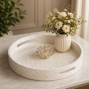 May include: A round white tray with a textured, iridescent shell-like finish and two integrated side handles. Inside the tray sits a small, clear glass lotus-shaped candle holder and a ribbed cream-colored vase filled with white flowers.