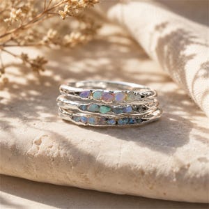 May include: A stack of three silver rings, each featuring a row of small, raw, iridescent opal stones embedded within the metal band. The silver has a textured, organic finish, and the opals display flashes of blue, green, and pink colors.