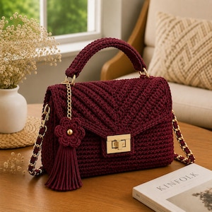 May include: A deep burgundy crochet handbag featuring a chevron pattern, a gold-tone metal turn-lock closure, and a matching crochet flower tassel charm. The bag includes a sturdy top handle and a gold chain strap woven with matching yarn.