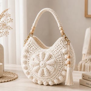 May include: A cream-colored crocheted handbag with a crescent shape, featuring a central raised flower motif and small pom-pom accents along the bottom edge. The bag includes a braided handle, light wood beads, and a matching tassel attachment.
