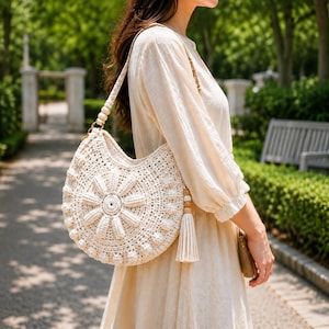 May include: A cream-colored crochet shoulder bag featuring a large, raised floral motif on the front. The bag has a crescent shape, a braided strap adorned with wooden beads, and a long, matching cream-colored tassel hanging from the side.