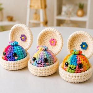 Può includere: Tre creature amigurumi all'uncinetto in conchiglie aperte color crema. Ogni creatura è rotonda, multicolore e presenta occhi neri e un sorriso ricamato. L'interno di ogni coperchio a conchiglia è decorato con un piccolo fiore all'uncinetto.