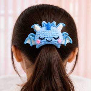 Könnte beinhalten: Ein hellblaues gehäkeltes Haaraccessoire in Drachenform mit weißen Innenflügeln, einem kleinen blauen Kamm und rosa runden Wangen. Der Drache hat geschlossene Augen und ein einfaches schwarzes gesticktes Lächeln, befestigt an einem dunkelbraunen Pferdeschwanz.