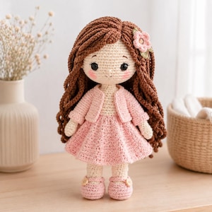 Peut inclure: Une poupée en crochet faite à la main avec de longs cheveux en laine brune ondulée, ornée d'une petite fleur rose. La poupée porte un gilet en crochet rose clair et une jupe plissée assortie, avec des chaussures roses à petits boutons en bois.