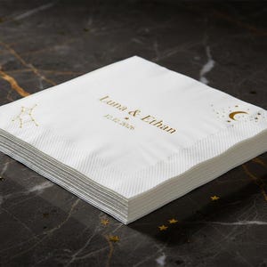 May include: A stack of white paper napkins featuring gold foil printing. The design includes the text Luna & Ethan, the date 12.12.2026, a constellation graphic on the left, and a crescent moon with small stars on the right side.