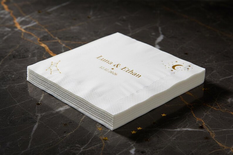 May include: A stack of white paper napkins featuring gold foil printing. The design includes the text Luna & Ethan, the date 12.12.2026, a constellation graphic on the left, and a crescent moon with small stars on the right side.