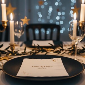 May include: A matte black dinner plate holds a white square napkin printed with the text Luna & Ethan and the date 12.12.2026, featuring a gold crescent moon and star design. The table setting includes silver cutlery, wine glasses, and candles.