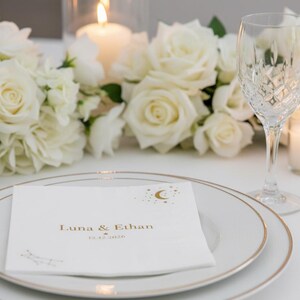 May include: A white paper napkin featuring gold foil text that reads Luna & Ethan with the date 12.12.2026 and a crescent moon and star graphic. The napkin is placed on a white plate with a gold rim, next to a crystal glass and white roses.