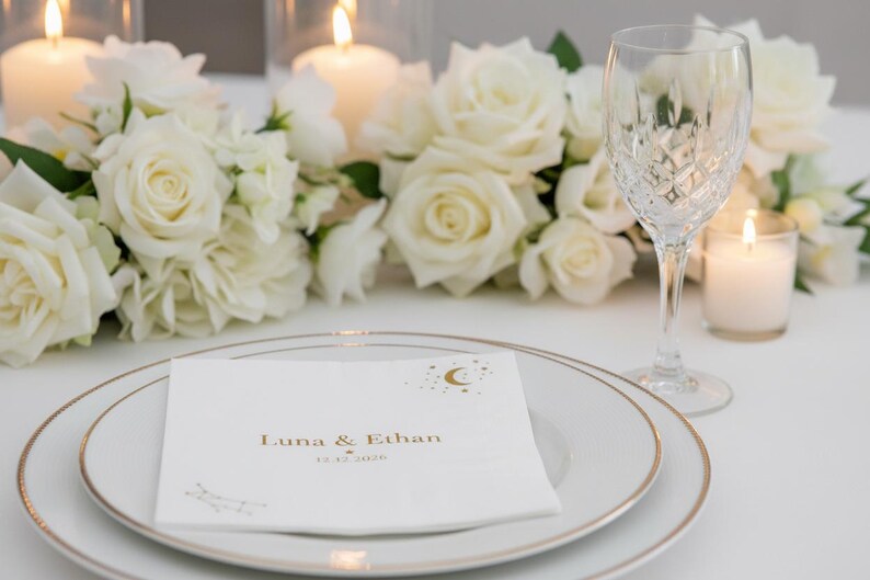 May include: A white paper napkin featuring gold foil text that reads Luna & Ethan with the date 12.12.2026 and a crescent moon and star graphic. The napkin is placed on a white plate with a gold rim, next to a crystal glass and white roses.