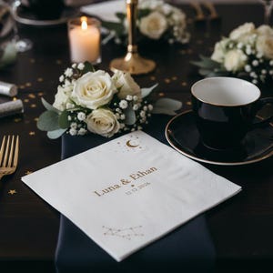 May include: A white square paper napkin featuring gold foil text that reads Luna & Ethan, the date 12.12.2026, a crescent moon, stars, and a constellation graphic. The napkin is placed on a dark table setting with gold cutlery and a black teacup.