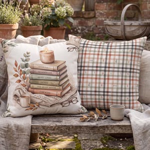 May include: Two square throw pillows. One pillow features a printed illustration of a stack of books titled Stories, Wanderlust, Quiet Lives, Home, The Quiet Hours, and Dreams, topped with a candle, a mug, and glasses. The second pillow has a plaid pattern.